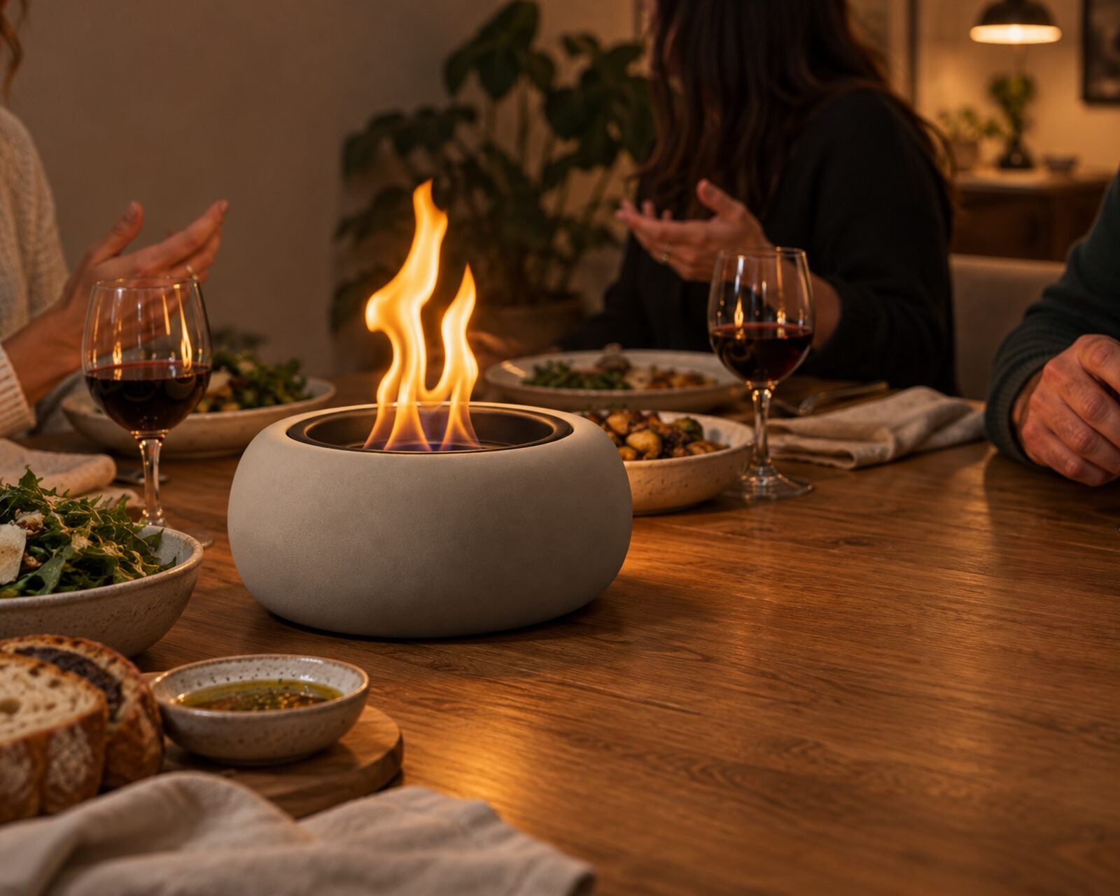 A tabletop fire bowl burning as the centerpiece of a dinner table indoors, friends sharing a meal around it