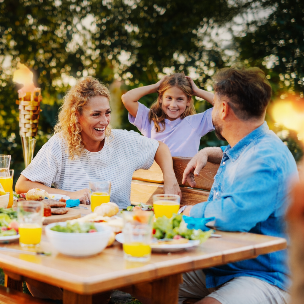 Family enjoying a meal outdoors with a wooden table and greenery in the background